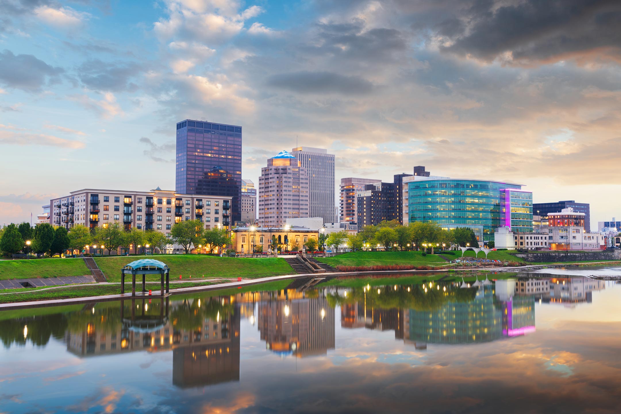Careers at ID CardSolutions | Join the Team - Dayton-riverfront-skyline City skyline at sunset with modern glass buildings along a calm river and a grassy riverside park, reflecting in the water.